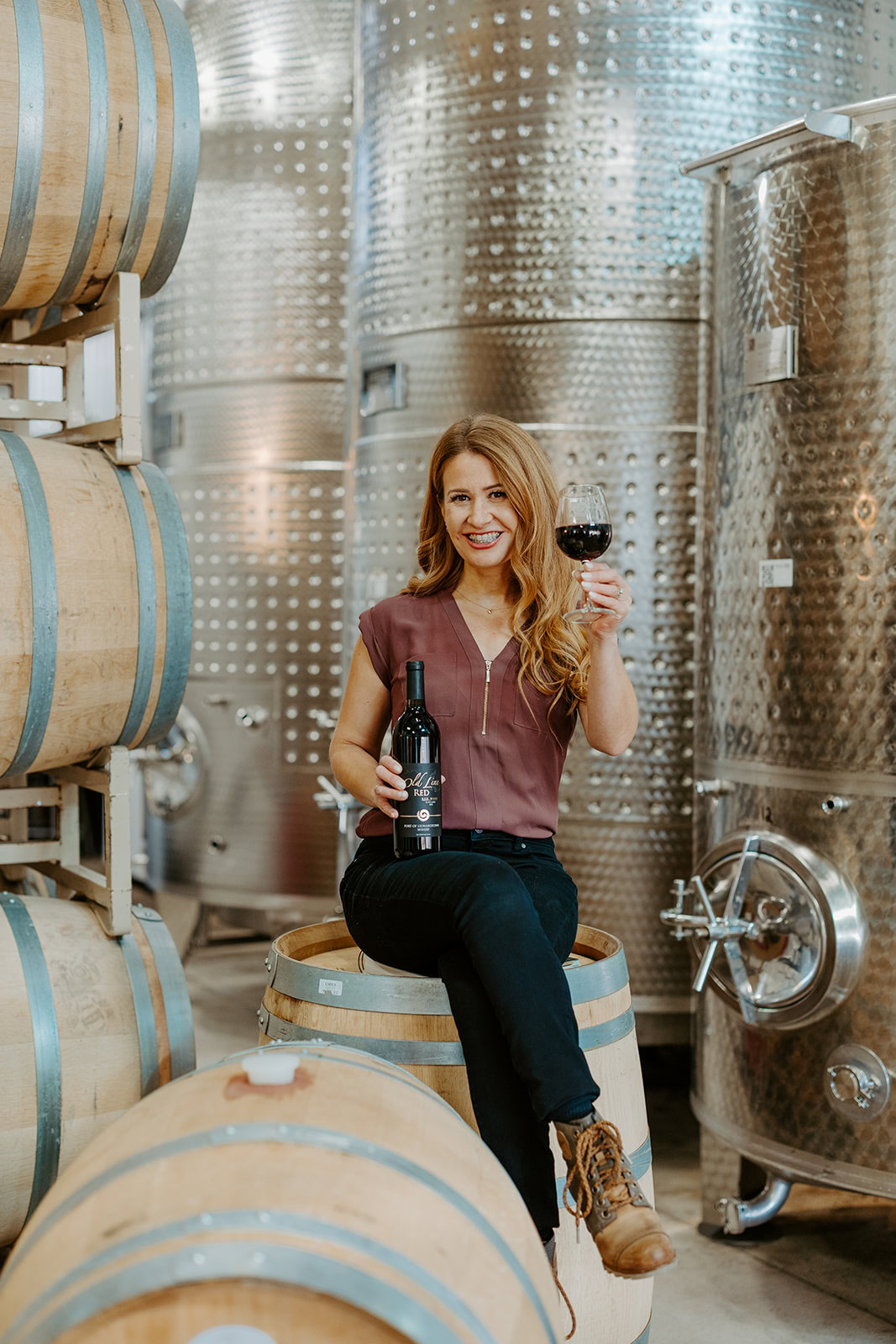 Woman holding a wine glass and bottle in a winery with stainless steel fermentation tanks.