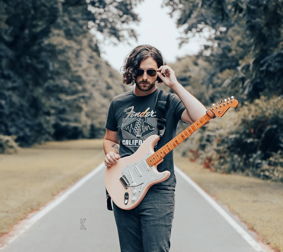 Man with sunglasses holding an electric guitar on a tree-lined road.